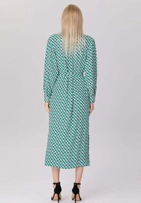 Femme aux longs cheveux blonds portant une robe midi à motifs géométriques teal et blancs et des sandales à talons hauts noires, se tenant de dos.