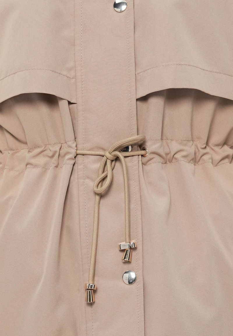 Beżowa kurtka z wcięciem w talii, z troczkami z metalowymi akcentami i zatrzaskami. Gładki materiał i luźny krój.