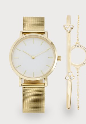 Montre dorée avec un bracelet en maille, dotée d'un cadran blanc et de marqueurs d'heure minimalistes. Inclut un bracelet doré avec trois accents en cristal.