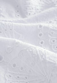 Tissu blanc brodé avec des motifs floraux complexes et des détails en œillets. Texture douce avec une apparence lisse et légère.