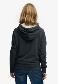 Mørkegrå hættetrøje med blødt hvidt foer, med snøre i hætten og ribbede manchetter og kant. Bæres med blå jeans.