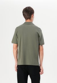 Moški, obrnjen stran, oblečen v olivno zeleno polo majico in črne hlače, stoji ob belem ozadju.