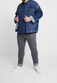 Jeansjacke in blauer Nuance, graue Ripp-Hosen und weiße Sneaker. Die Jacke verfügt über zwei Fronttaschen und umgekrempelte Ärmelsäume.