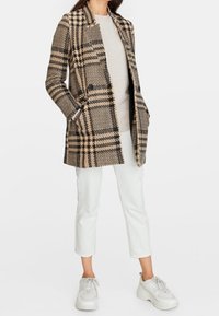 Nainen, jolla on päällään beige ja musta ruudullinen takki, beige villapaita, valkoiset lyhyet housut ja valkoiset korkeat lenkkarit, kädet takin taskuissa.