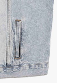 Jasnoniebieska kurtka dżinsowa z wyraźną kieszenią, widocznym przeszyciem oraz gładką fakturą z delikatnymi efektami wyblaknięcia.