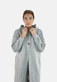 Hellgrauer, gestrickter langer Cardigan mit Kapuze, ausgestattet mit einem strukturierten Muster, zwei Knöpfen und gerippten Bündchen. Weicher Stoff mit lockerer Passform.