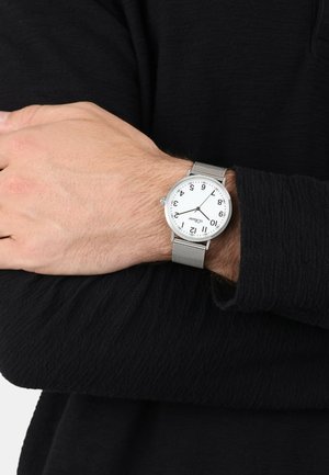 Handgelenk mit einer silbernen Mesh-Armbanduhr, die 10:10 anzeigt, vor dem Hintergrund eines schwarzen, langärmeligen strukturierten Hemdes.