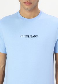 Hele sinine puuvillane T-särk, millel on ümmargune kaelus ja must "GUESS JEANS" logo, mis on tikkimistööna rindkerele kantud. Sile tekstuur, lühikesed varrukad.