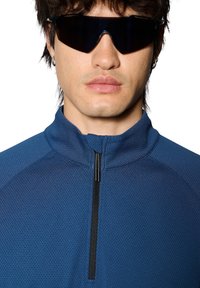 Blå zip-up tröja i ett strukturerat tyg, med hög krage och en svart dragkedja. Solglasögon med mörka linser fulländar looken.