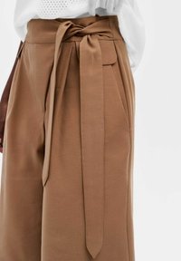 Bruine hoge taillebroek met een stoffen riem die om de taille is geknoopt, voorzien van zijzakken en een gladde, gestructureerde afwerking.