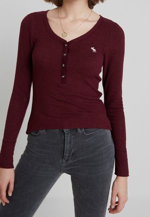 Mujer con una camiseta Henley ajustada de manga larga y canalé en color burdeos con botones, y jeans gris oscuro, de pie frente a un fondo liso.
