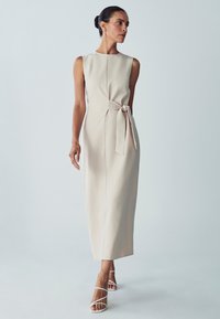 Beige midi dress with a fitted silhouette, round neckline, and a side tie accent. Smooth fabric and sleeveless design. White strappy heels.