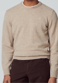 Beige knit sweater with a crew neckline and ribbed cuffs. Features a small logo on the chest. Worn over dark brown trousers.