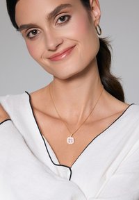 Złoty naszyjnik z kwadratowym zawieszką, ozdobiony wieloma różowymi i przezroczystymi kamieniami szlachetnymi. Noszony na białej bluzce z czarnym wykończeniem. Gładka faktura.