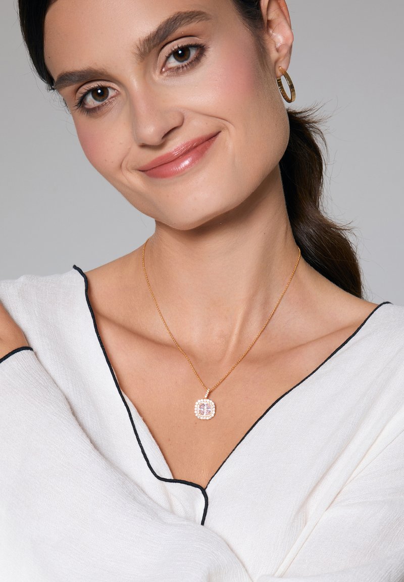 Złoty naszyjnik z kwadratowym zawieszką, ozdobiony wieloma różowymi i przezroczystymi kamieniami szlachetnymi. Noszony na białej bluzce z czarnym wykończeniem. Gładka faktura.