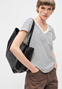 Μαύρη δερμάτινη τσάντα τύπου tote με λεία υφή, με διπλούς ιμάντες ώμου και μινιμαλιστικό σχέδιο, συνδυασμένη με ένα ριγέ μπλουζάκι V-neck.