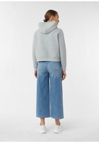 Kobieta z brązowymi włosami w niskim kokie, ubrana w szary hoodie, szerokie, ścięte niebieskie dżinsy oraz białe sneakersy, stojąca na tle jednolitego białego tła.
