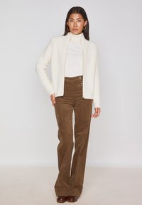 Cardigan crema con finitura testurizzata, chiusura con zip, indossato sopra un dolcevita bianco e dei pantaloni a zampa d'elefante in corduroy marroni a vita alta.