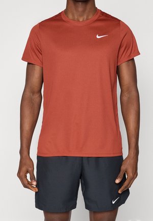 Mand iført rustfarvet Nike T-shirt med korte ærmer og sorte Nike-shorts, der står foran en ensartet lys baggrund.