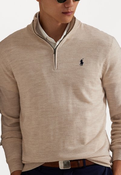 Pull beige à fermeture éclair quart avec des manches longues, texture tricotée et accent de logo foncé. Porté par-dessus une chemise à col et associé à une ceinture marron.