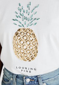 T-shirt bianco con un design di ananas ricoperto di paillettes dorate e decorazioni turchesi sopra, e il testo "LOOKING PINE" sotto.