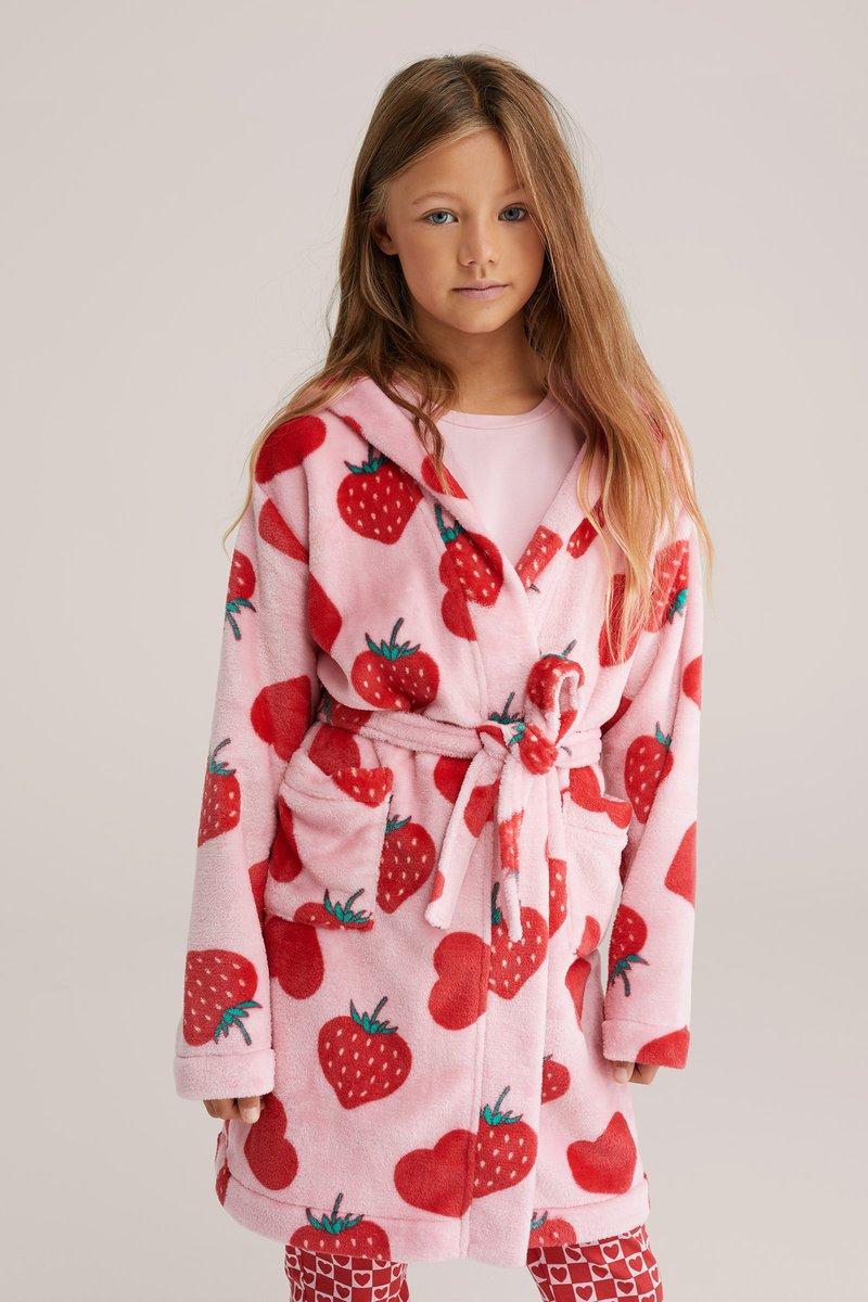 Fille aux longs cheveux portant un peignoir rose orné de motifs de fraises rouges et de cœurs, noué à la taille, debout devant un fond uni.