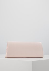 Jasnoróżowa satynowa kopertówka w kształcie trójkątnego pryzmatu leżąca na białej powierzchni na tle jednolitego szarego tła.