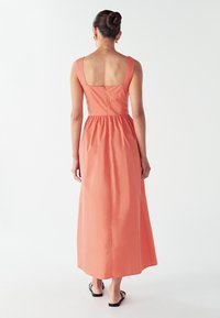 Žena s punđom koja nosi bezrukavnu koraljnu maksi haljinu i crne sandale, stoji pred bijelom pozadinom.