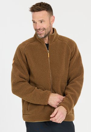 Mann trägt eine braune Fleecejacke mit Reißverschluss, richtet die Manschette, lächelt und schaut seitlich vor einem weißen Hintergrund.