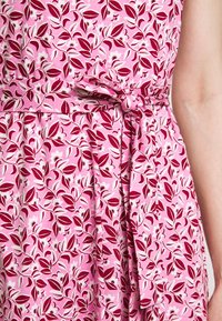 Robe rose avec motif floral rouge et blanc, ceinture en tissu nouée à la taille et design sans manches.