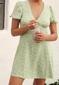 Robe mini verte à fleurs avec de courtes manches bouffantes, décolleté en V et texture douce. Le mannequin tient une bouteille en verre avec une fermeture à fil.