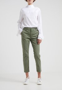 Person som bär en vit långärmad skjorta, olivgröna byxor med händerna i fickorna, och vita sneakers på en enhetlig ljus bakgrund.