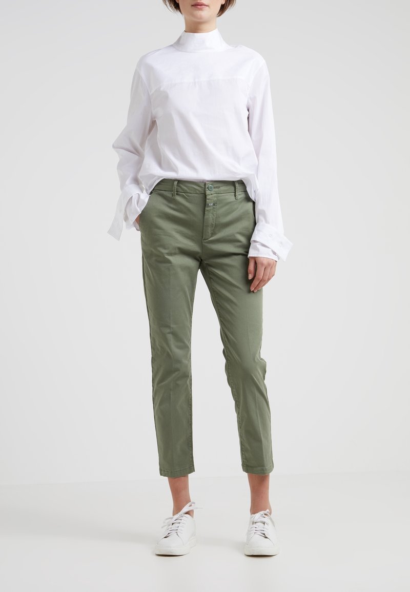 Person som bär en vit långärmad skjorta, olivgröna byxor med händerna i fickorna, och vita sneakers på en enhetlig ljus bakgrund.