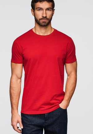 Viiksekäs mies, joka pukee yksivärisen punaisen lyhythihaisen t-paidan ja tummat farkut, seisoo käsi yhdessä taskussa vaaleaa taustaa vasten.