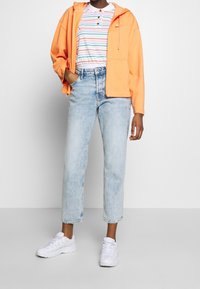 Oranssi vetoketjuhuppari valkoisen ja raidallisen polo-paidan päällä, yhdistettynä vaaleansinisillä suoralinjaisilla farkuilla ja valkoisilla tennareilla.