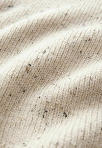 Tissu tricoté de couleur crème avec une texture côtelée et de petites taches noires dispersées sur le matériau.