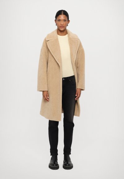 Beige faux fur coat with a wide collar and open front, paired with a light cream top and black skinny jeans. Black ankle boots complete the look.