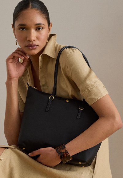 Femme en robe chemise beige tenant un sac fourre-tout en cuir noir avec des accents dorés, portant un bracelet en écaille de tortue et de petites boucles d'oreilles gouttes en or.