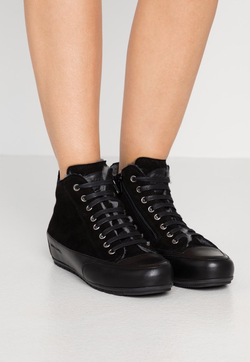 Baskets montantes noires avec fermeture éclair latérale et design lacé portées sur des jambes nues, présentant des matériaux en daim et cuir.