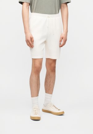 Hombre que lleva pantalones cortos crema acanalados con cordón, zapatillas beige con suelas de goma y una camisa de manga corta verde apagado, de pie sobre un fondo blanco.