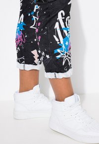 Pantalons noirs avec des motifs de graffiti colorés en bleu, rose et blanc, présentant un ourlet effiloché et associés à des baskets montantes blanches.