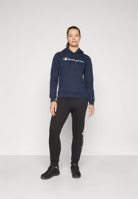 Tumesinine kapuutsärk valge "Champion" logo ja taskuga, koos mustade joggeritega külglogo printimisega ning mustade spordijalanõudega.
