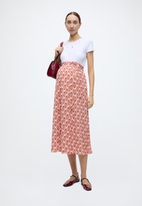 Haut blanc à manches courtes côtelé associé à une longue jupe rouge à motifs géométriques, accompagné d'un sac à main rouge et de chaussures assorties.