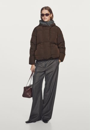 Bruine pufferjas met capuchon en doorgestikt ontwerp, gecombineerd met grijze wijde broekspijpen en een bordeauxrode handtas vasthoudend.