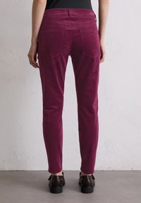 Bordeauxrote Cordhosen im Slim-Fit-Stil, mit Gesäßtaschen und Gürtelschlaufen, kombiniert mit schwarzem Schuhwerk auf einem hellgrauen Boden.
