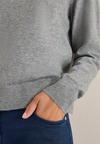 Maglione grigio realizzato in morbido materiale con orlo e polsini a costine, abbinato a jeans blu scuro con dettaglio a bottoni sulla tasca.