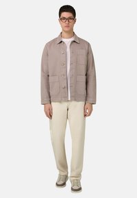 Hellgraue Jacke mit Knopfleiste, zwei Brusttaschen, getragen über einem weißen Hemd, kombiniert mit cremefarbenen Hosen und grauen Turnschuhen.