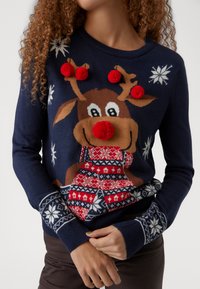 Pulover bleumarin cu un design de ren, decorat cu pompoane roșii și fulgi de zăpadă. Modelul tricotat include accente geometrice în roșu și bleumarin.