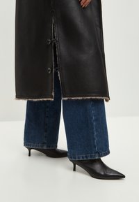 Musta nahkatakki, jossa on karvavuori, napitettava suljenta ja sivuhalkiot. Yhdistetty leveälahkeisiin sinisiin farkkuihin ja kärjellisiin mustiin korkokenkiin.