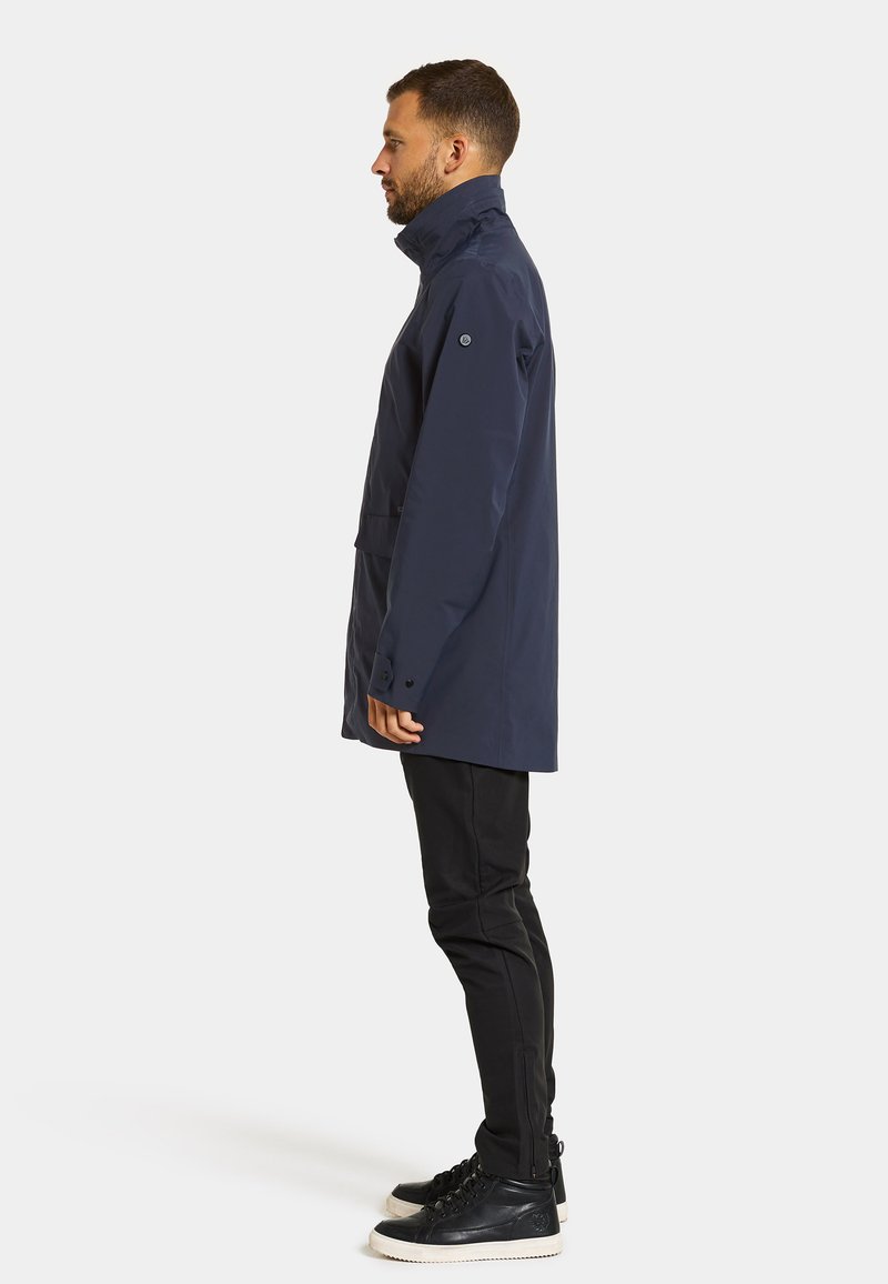 Marineblaue wasserdichte Jacke mit hohem Kragen, langen Ärmeln und Fronttasche, kombiniert mit schwarzen Slim-Fit-Hosen und schwarzen High-Top-Sneakern.